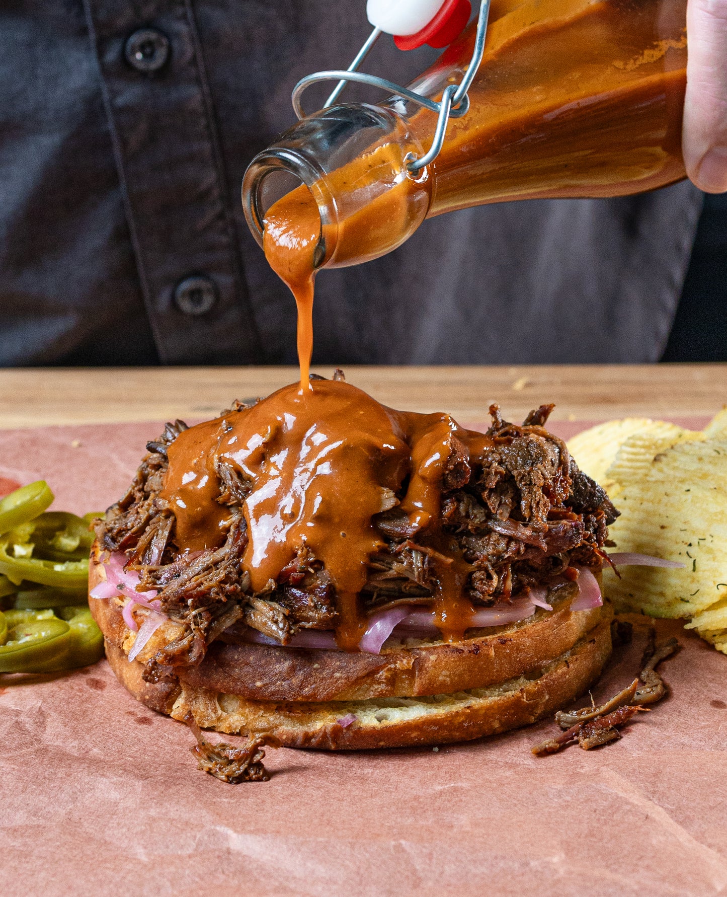 Braised meat sandwich being drizzled with barbecue sauce on a wooden surface.