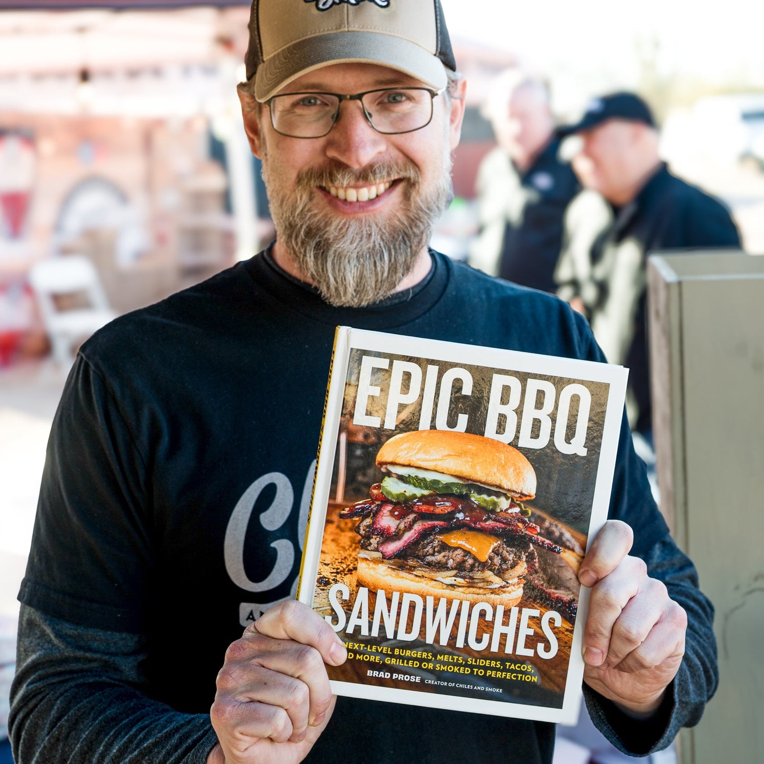 Brad Prose holding cookbook titled 'Epic BBQ Sandwiches' at an outdoor event.