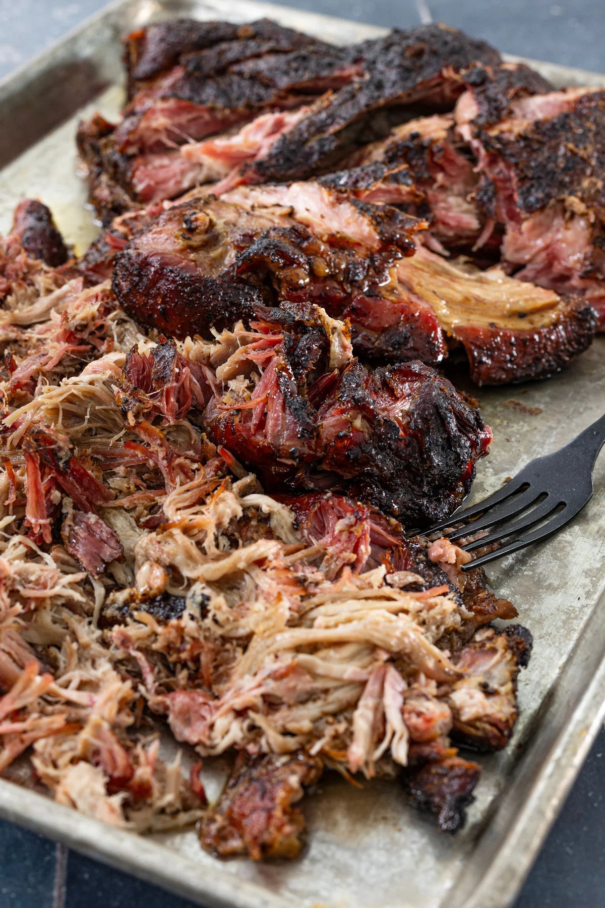 Close-up of pulled barbecue meat on a metal baking sheet with a black fork, showing dark caramelized exterior and shredded interior.