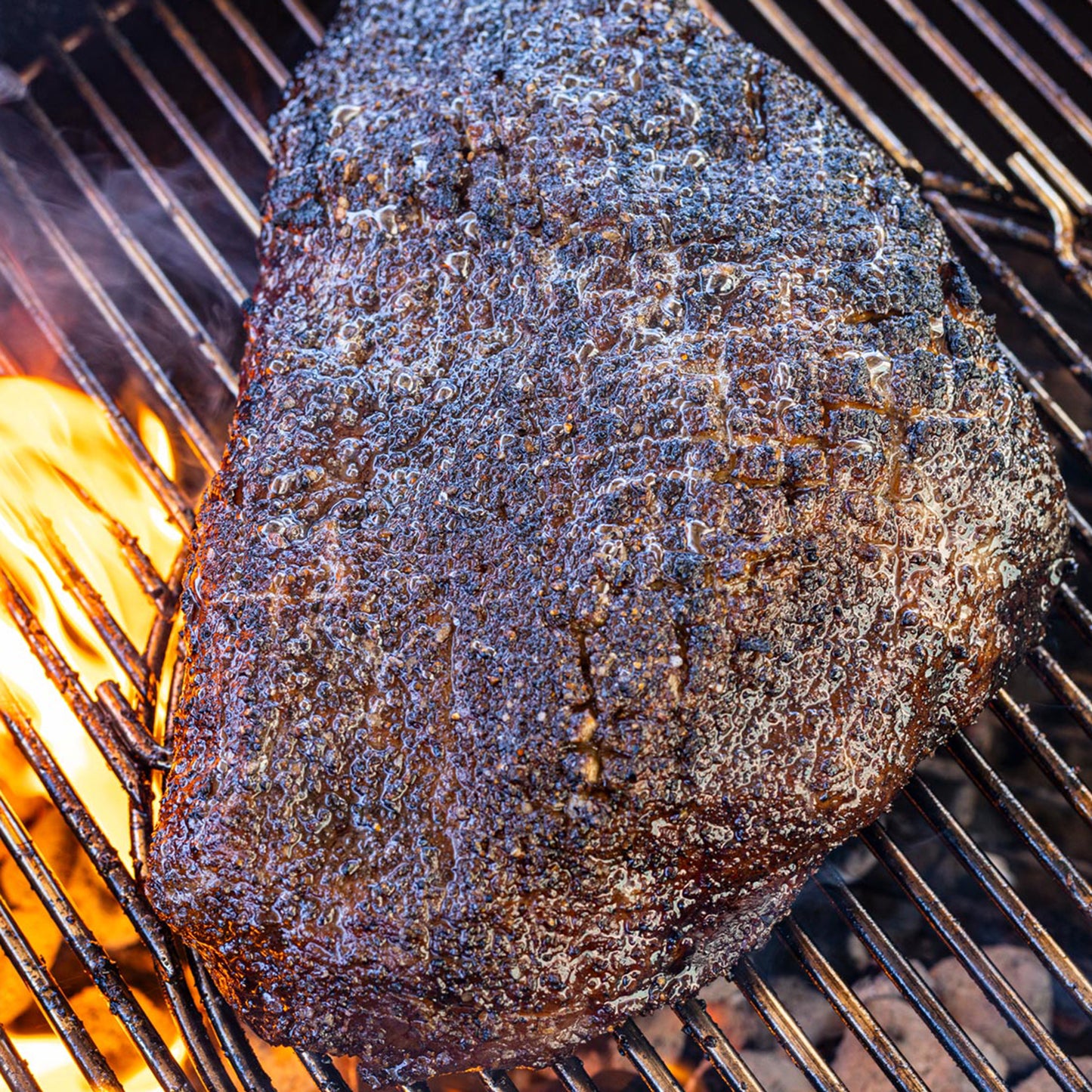 Seasoned steak with dark peppery bark cooking over fire on a charcoal grill