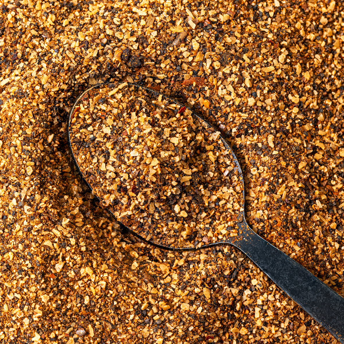 Macro close-up of Chiles and Smoke Canyon Crust Beef Seasoning, a coarse salt and pepper rub, on a black spoon
