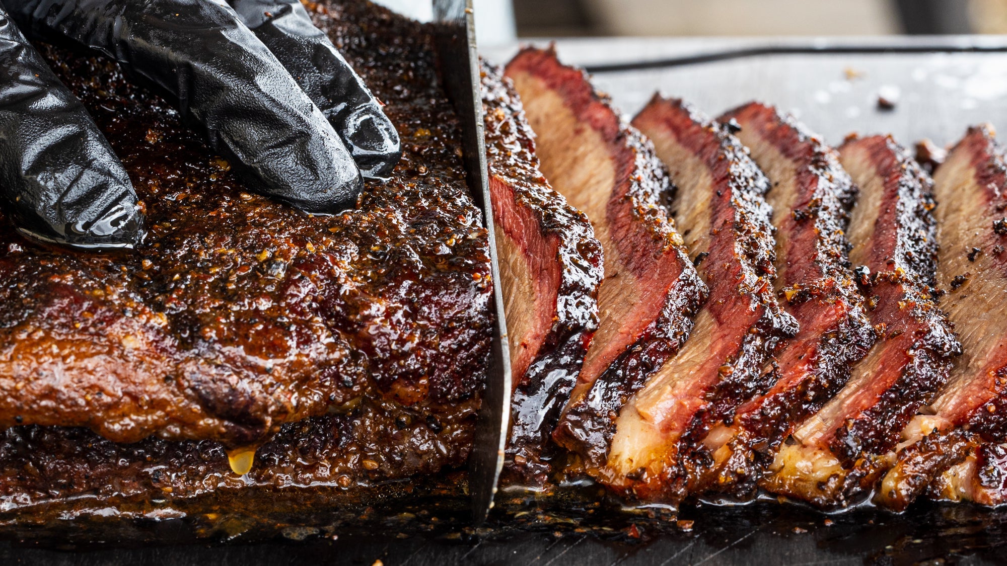 Sliced smoked brisket with a knife and gloved hand on a cutting board.